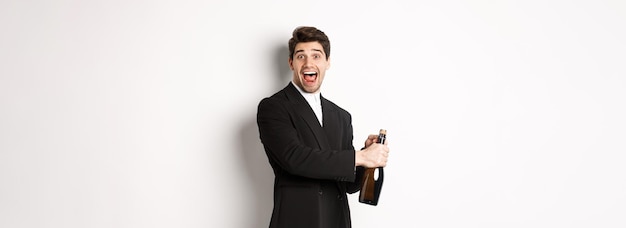 Free photo portrait of attractive man in black suit winking at camera and opening bottle of champagne celebrati