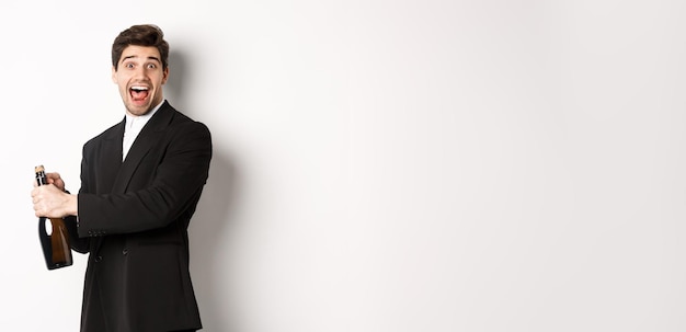 Free photo portrait of attractive man in black suit winking at camera and opening bottle of champagne celebrati