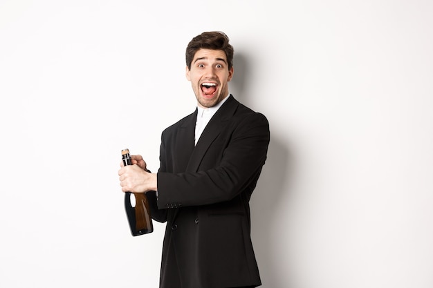 Portrait of attractive man in black suit, winking at camera and opening bottle of champagne, celebrating new year, standing against white background.