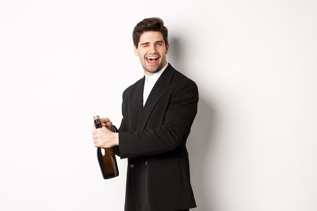 Портрет привлекательного мужчины в черном костюме, подмигивающего в камеру и открывающего бутылку шампанского, празднующего Новый год, стоя на белом фоне.