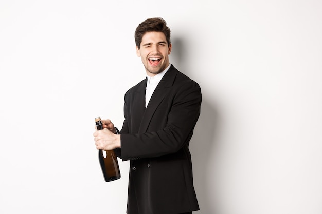 Portrait of attractive man in black suit, winking at camera and opening bottle of champagne, celebrating new year, standing against white background.