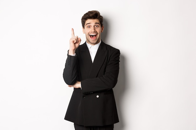 Free photo portrait of attractive man in suit, having an idea, standing excited and raising one finger to tell suggestion, think-up solution, standing over white background