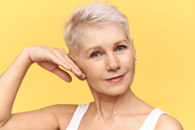 Portrait of attractive middle aged Caucasian woman in tank top touching cheek, applying anti wrinkle moisturizing cream, looking at camera with smile.