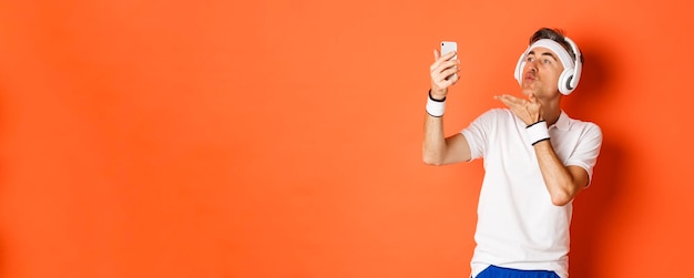 Portrait of attractive middleaged man in gym uniform wearing headphones sending air kiss at mobile p