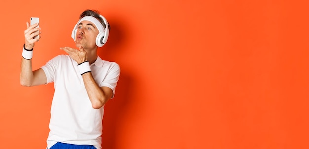 Free photo portrait of attractive middleaged man in gym uniform wearing headphones sending air kiss at mobile p