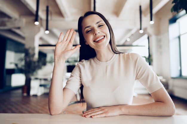 Портрет привлекательной улыбающейся деловой женщины, махающей рукой, смотрящей в камеру