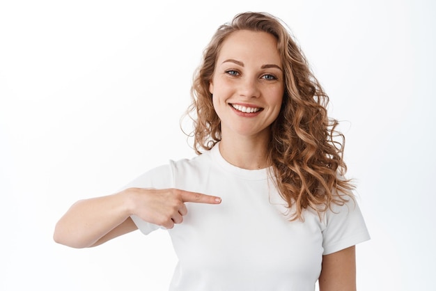 Portrait of attractive smiling girl showing your logo pointing finger aside at copyspace and looking satisfied recommending advertisement white background