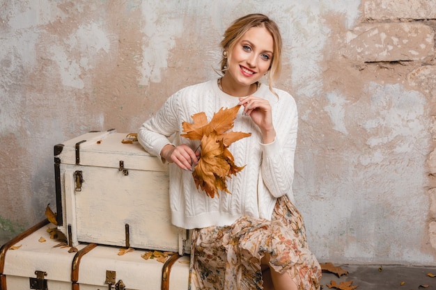 Portrait of attractive smiling stylish blonde woman in white knitted sweater sitting on suitcases in street against vintage wall