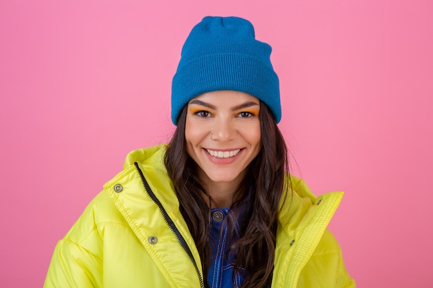 Portrait of attractive smiling stylish woman posing on pink wall in colorful winter down jacket of yellow color, wearing blue knitted hat, dressed in warm clothes, fashion trend