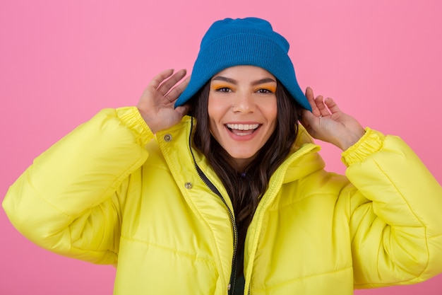 Portrait of attractive smiling stylish woman posing on pink wall in colorful winter down jacket of yellow color, wearing blue knitted hat, dressed in warm clothes, fashion trend