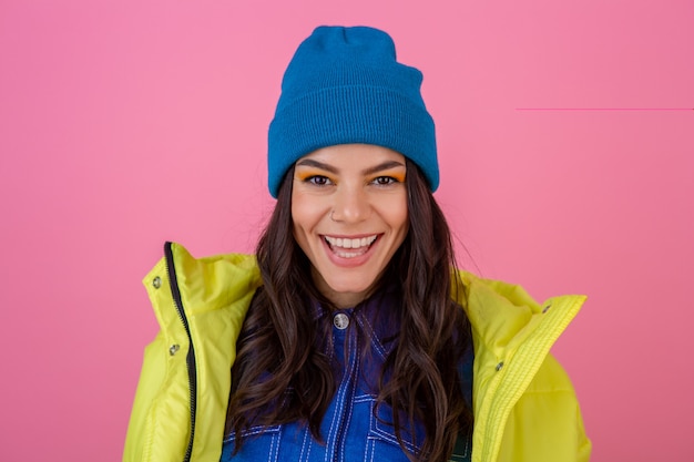 冬のファッションでポーズをとる魅力的な笑顔のスタイリッシュな女性の肖像画は、暖かい服を着て、青いニット帽を身に着けている明るいネオン黄色のジャケットのピンクの壁に見えます