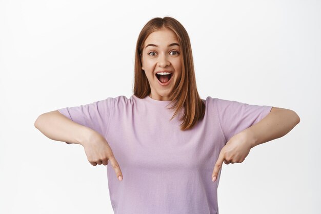 魅力的な笑顔の女性が指を下に向け、興奮して笑って、広告、ロゴ、またはバナーを表示し、白い背景のTシャツに立っている肖像画。