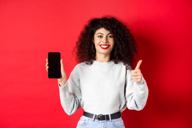Portrait of attractive smiling woman with curly hair, showing empty mobile phone screen and thumb-up, recommending online promo, standing on red background.