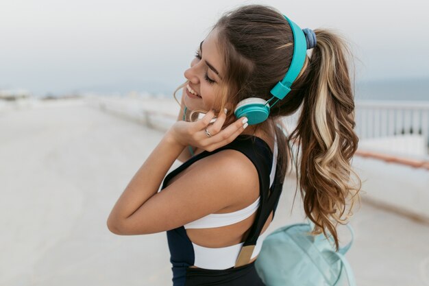 Portrait attractive sportswoman with long curly hair in headphones walking in the early morning on seafront. Enjoying training outside, cheerful mood, lovely music, smiling with closed eyes