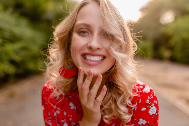 Portrait of attractive stylish blond smiling woman in red blouse summer fashion outfit in park boho style wearing earrings smiling 