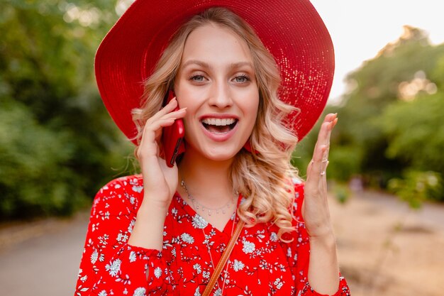 Портрет привлекательной стильной блондинки, улыбающейся женщины в соломенной красной шляпе и блузке, летней модной одежде, разговаривает по телефону с положительными эмоциями жестов