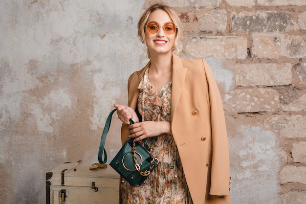 Portrait of attractive stylish blonde woman in beige coat walking in street against vintage wall