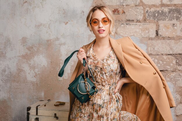 Portrait of attractive stylish blonde woman in beige coat walking in street against vintage wall