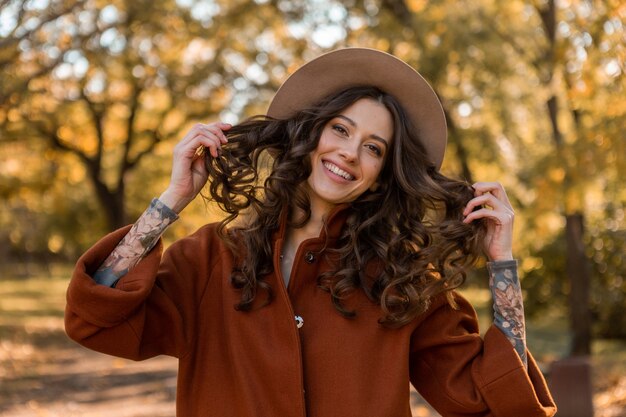 Portrait of attractive stylish smiling woman with long curly hair walking in park dressed in warm brown coat autumn trendy fashion, street style wearing hat