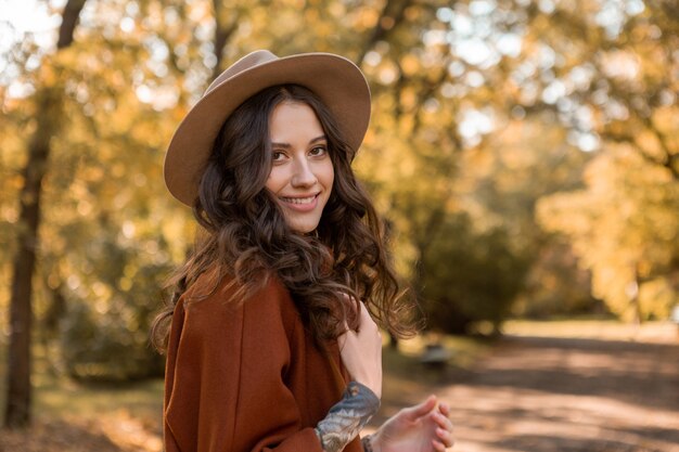 Portrait of attractive stylish smiling woman with long curly hair walking in park dressed in warm brown coat autumn trendy fashion, street style wearing hat