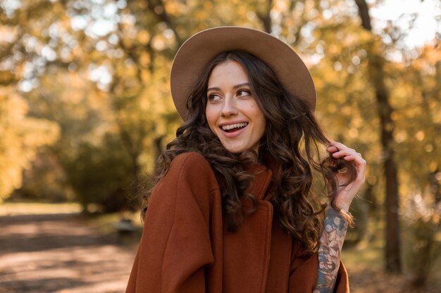 Portrait of attractive stylish smiling woman with long curly hair walking in park dressed in warm brown coat autumn trendy fashion, street style wearing hat