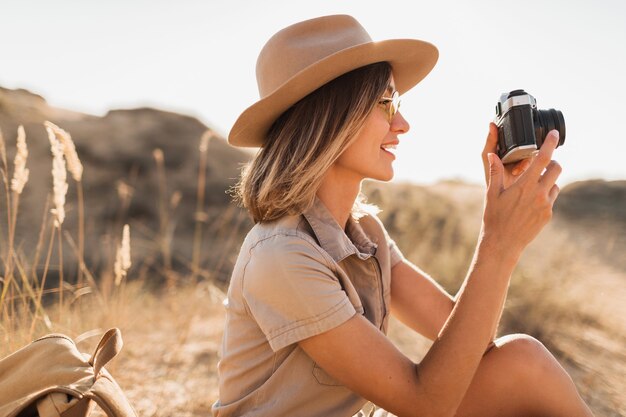 Portrait of attractive stylish young woman in khaki dress in desert, traveling in Africa on safari, wearing hat, taking photo on vintage camera