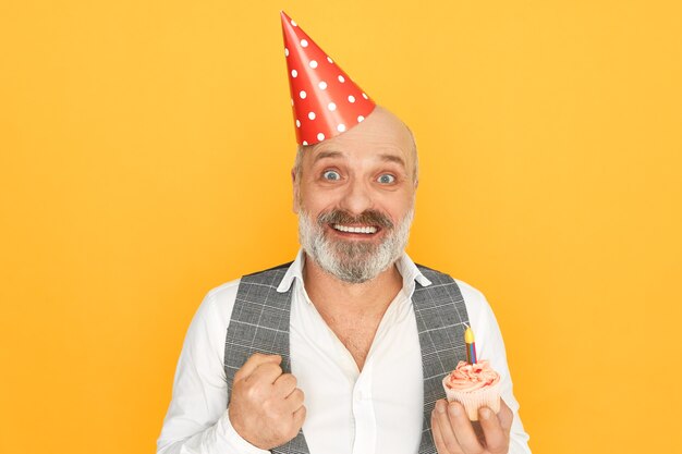 Portrait of attractive successful senior bearded businessman wearing cone hat expressing excitement, enjoying birthday party, holding cupcake with one candle