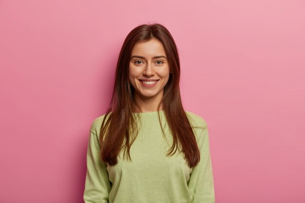 Портрет привлекательной женщины со здоровой кожей, зубастой улыбкой, прямым взглядом, зеленым джемпером, длинными прямыми волосами, позирует на фоне розовой пастельной стены. Концепция выражения лица