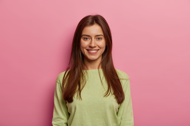 Portrait of attractive woman has healthy skin, has toothy smile, looks directly , wears green jumper, has long straight hair, poses against pink pastel wall. Face expressions concept