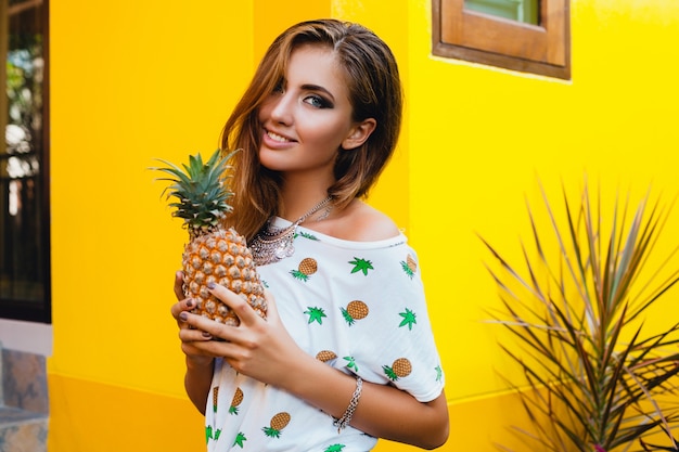 Portrait of attractive woman in pinted t-shirt on summer vacation holding pineapple, fruit diet detox, tanned skin, bright yellow background