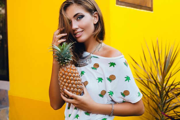 Portrait of attractive woman in pinted t-shirt on summer vacation holding pineapple, fruit diet detox, tanned skin, bright yellow background