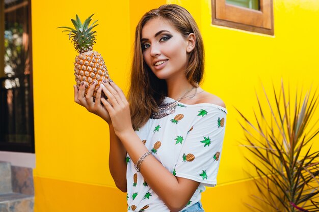 Portrait of attractive woman in pinted t-shirt on summer vacation holding pineapple, fruit diet detox, tanned skin, bright yellow background