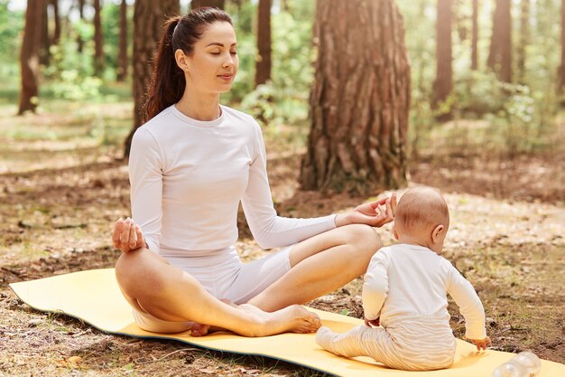 Portrait of attractive woman sitting in lotus pose and meditating while her infant daughter