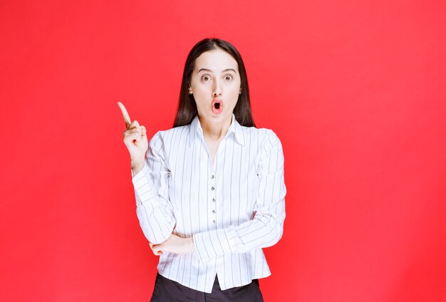 Portrait of attractive woman standing and pointing fingers up against red wall. 