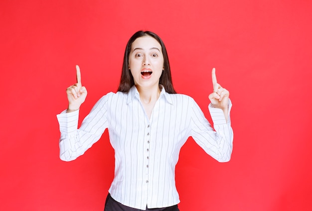 Portrait of attractive woman standing and pointing fingers up against red wall. 