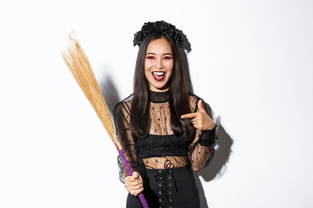 Portrait of attractive woman in witch costume celebrating halloween, pointing at broom, standing over white background.