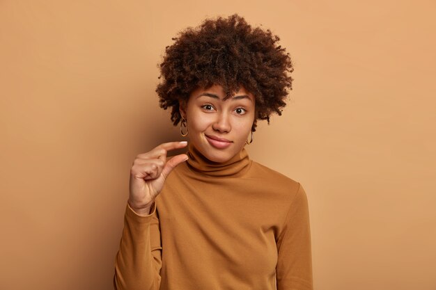 Portrait of attractive woman with curly Afro hair, shapes tiny and small thing, talks about size, dressed in casual brown jumper