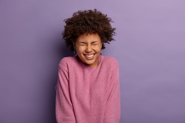 Foto gratuita ritratto di donna attraente con acconciatura riccia, scherza felicemente, sente una storia divertente e divertente, indossa un maglione casual