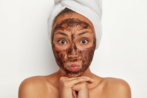 Portrait of attractive woman with surpised expression, has coffee scrub around face, cleanses from pores, removes dead cells, chooses mask to suit her skin, stands topless after shower, towel on head