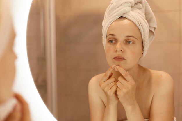 Portrait of attractive woman wrapped in white towel standing with bare shoulders in bathroom and looking for or squeezing acne on chin, having sad expression.