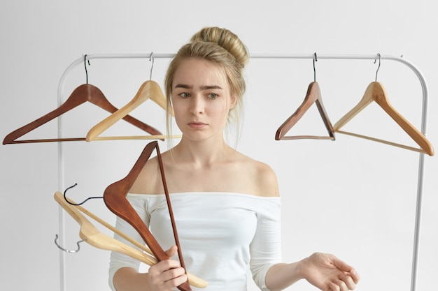 portrait of attractive worried woman with hair bun posing against white wall, holding racks in her hands while cleaning her wardrobe, getting rid off useless clothes. Nothing to wear