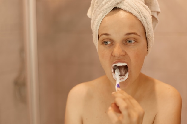 口を大きく開けて歯を磨き、自宅のバスルームで衛生管理を行い、タオルに包まれた魅力的な若い成人女性の肖像画。