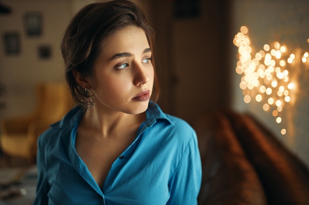 Free photo portrait of attractive young caucasian woman with gathered hair posing in stylish apartment with blurred lights in background, having upset bored facial expression