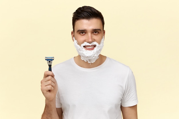 Portrait of attractive young dark haired guy in casual t-shirt posing with shaving cream on his face, holding disposable razor