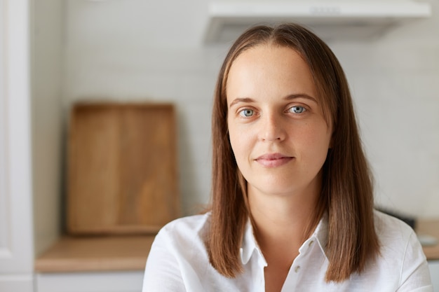 明るい部屋で自宅で魅力的な若い黒髪の女性の肖像画、穏やかな表情でカメラを見て、白いシャツを着て、屋内ショットの美しい女性。