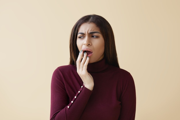 Portrait of attractive young dark skinned female having bored facial expression, looking away, covering mouth while yawning, feeling tired during working day at office. Human gestures and signs