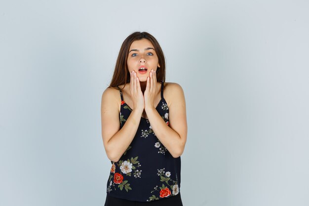 Portrait of attractive young lady holding hands near open mouth in blouse and looking surprised front view