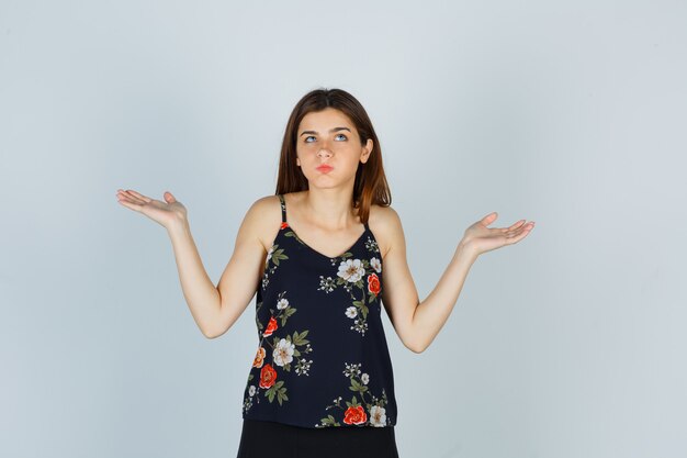 Portrait of attractive young lady showing helpless gesture, blowing cheeks in blouse and looking confused front view