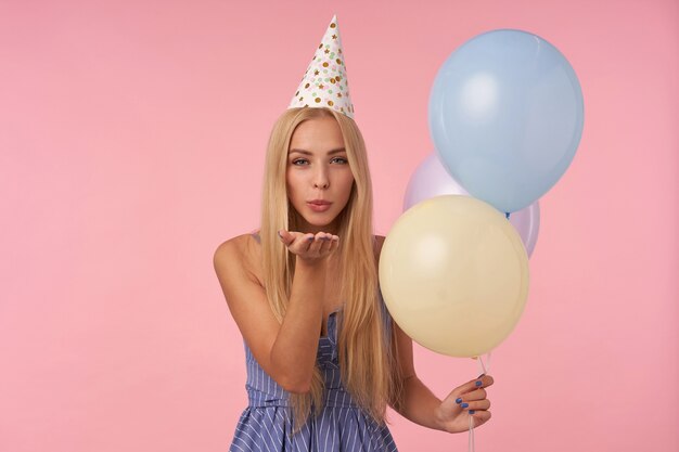 Portrait of attractive young long haired woman celebrating birthday with multicolored air balloons, looking at camera positively and blowing air kiss with pursed lips