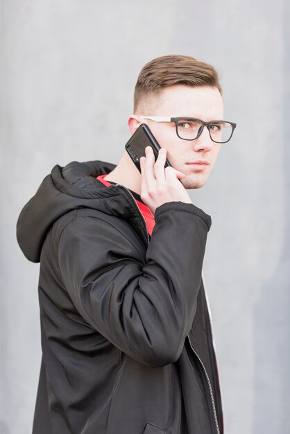 Portrait of an attractive young man with eyeglasses talking on mobile phone against grey backdrop